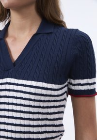 Polo a righe navy e bianche in maglia con texture a trecce, caratterizzato da un colletto a V e bordi rossi a contrasto sulle maniche.
