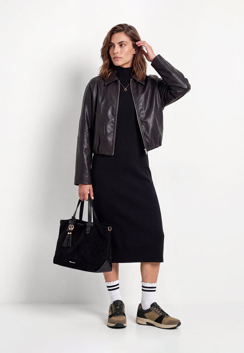 Femme portant une veste en cuir noire sur une robe noire, tenant un sac à main noir, avec des chaussettes blanches et des baskets marron, debout devant un fond blanc.