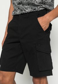 Schwarze Cargo-Shorts aus Baumwolle mit Seitentaschen und lockerer Passform. Der Stoff hat eine glatte Textur mit sichtbaren Nähdetails.