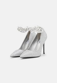 Escarpins argentés à bouts pointus, ornés de détails ressemblant à des cristaux et d'une bride de cheville décorative avec une chaîne. Talons aiguilles noirs.