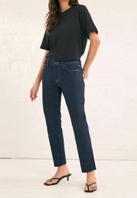 Vaqueros de denim azul oscuro con un ajuste recto y ceñido, cinco bolsillos y costuras en contraste. Combinados con una camisa negra de manga corta y sandalias planas.