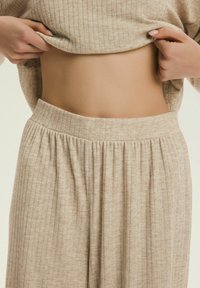 Ruce zvedají béžový žebrovaný pletený svetr, odhalující holé břicho nad odpovídajícími žebrovanými pletenými kalhotami.