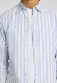 Vaaleansininen ja valkoinen raidallinen napapaidan, joka on valmistettu puuvillasta ja jossa on klassinen kaulus ja helmimäiset napit, käytetään valkoisen t-paidan päällä.