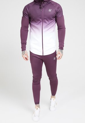 Mand iført lilla og hvid gradient lynlåstrøje og matchende lilla joggingbukser med hvide sneakers, der står mod en ensfarvet baggrund.