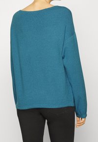 Pull en tricot turquoise avec un large col, des manches longues et une coupe décontractée. Texture lisse et ourlet légèrement lâche à la taille.