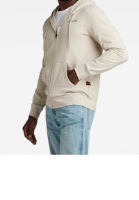 Felpa con zip beige realizzata in tessuto morbido, con tasca frontale e cappuccio con coulisse. Include dettagli con logo sul petto.