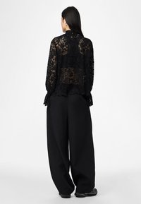 Top de renda preta com mangas compridas e bainha ondulada, combinado com calças pretas largas. Padrão floral detalhado. Tecido de renda texturizado.