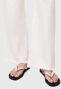 Infradito nere con cinturini in argento, indossate con pantaloni bianchi a gamba larga con orli grezzi, che mostrano una texture liscia e un design casual.