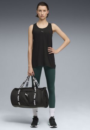 Top sin mangas negro, leggins verdes, bolsa de deporte negra con marca blanca. Modelo de pie, con las manos en las caderas, usando zapatillas negras y calcetines blancos.