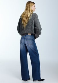 Széles szárú kék farmernadrág szürke kötött pulóverrel párosítva. A pulóver mintázott textúrával rendelkezik. A modell fekete magassarkút visel.