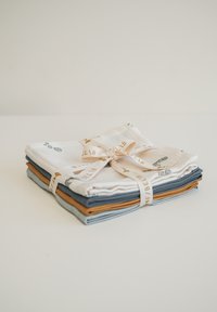 Sada čtyř bavlněných ubrousků složených na sebe; barvy zahrnují bílou s potisky, modrou, šedou a rezavě oranžovou. Přivázané krémovou stuhou.