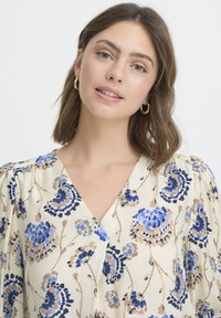 Bluse mit Blumenmuster, die blaue und beige Blumen auf einem hellen cremefarbenen Hintergrund zeigt, mit V-Ausschnitt und Puffärmeln.
