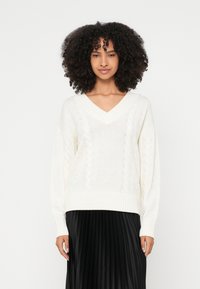 Maglione bianco lavorato a maglia con scollo a V, caratterizzato da motivi a trecce e polsini a coste, abbinato a una gonna nera plissettata.