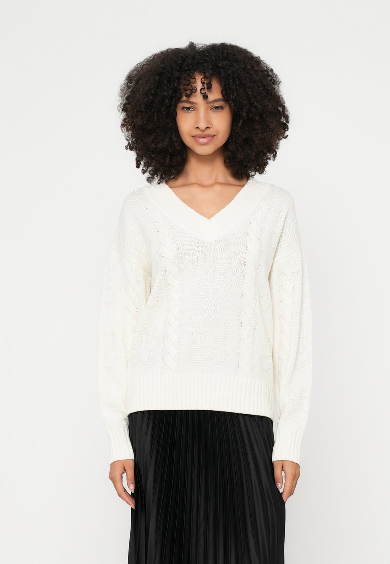 Maglione bianco lavorato a maglia con scollo a V, caratterizzato da motivi a trecce e polsini a coste, abbinato a una gonna nera plissettata.