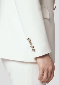 Biała marynarka w garniturze z rękawem ozdobionym czterema złotymi guzikami, uszyta z teksturowanej tkaniny, o prostym kroju.