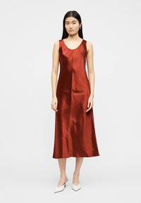 Fiatal nő áll egy egyszerű világosszürke háttér előtt, rozsdabarna színű, ujjatlan szatén midi ruhát és fehér hegyes orrú magassarkút visel.