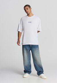 T-shirt bianco in cotone con maniche corte, con la stampa "Los Angeles" in blu. Abbinato a jeans di denim chiaro e larghi e sneakers bianche.