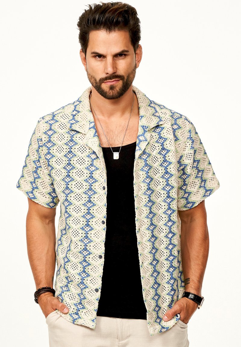 Kurzarmhemd in hellbeige mit blauen und grünen Zickzack-Mustern, aus strukturiertem Stoff. Schwarzes Tanktop darunter, kombiniert mit Accessoires.