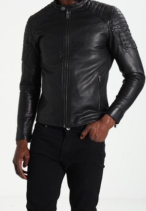 Homme portant une veste en cuir noire ajustée avec des détails matelassés aux épaules et un pantalon noir, une main dans la poche.