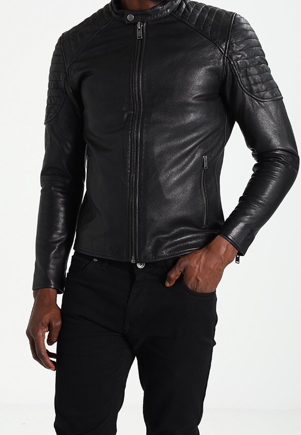 Homme portant une veste en cuir noire ajustée avec des détails matelassés aux épaules et un pantalon noir, une main dans la poche.