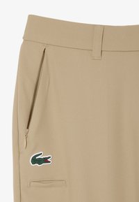 Pantaloni beige in tessuto liscio con tasca laterale con cerniera e logo coccodrillo ricamato bianco. Design classico con vestibilità su misura.