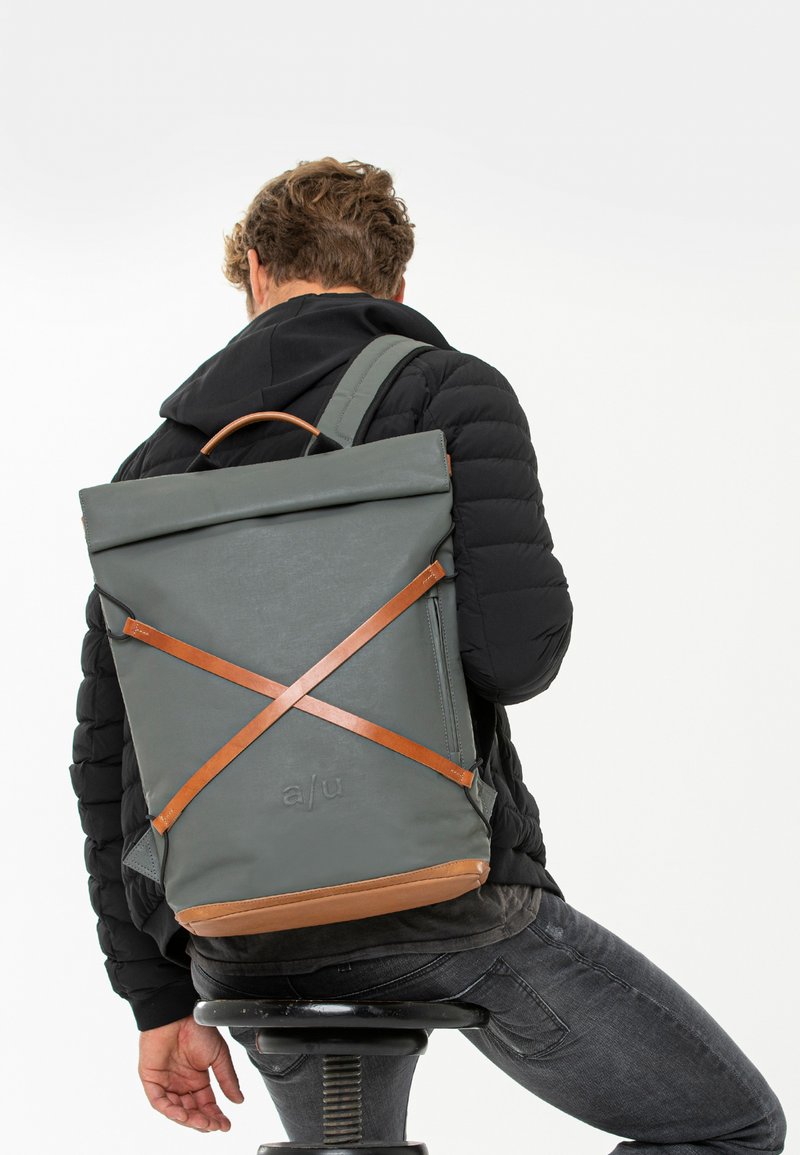 Grauer wasserdichter Rucksack mit braunen Lederakzenten und überkreuztem Riemen, ausgestattet mit einem Roll-Top-Design und einem Logo am Boden.