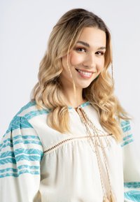 Blusa bianca con motivi geometrici blu sulle maniche, dotata di un colletto con cordino decorativo e tessuto testurizzato. Capelli acconciati in morbide onde.