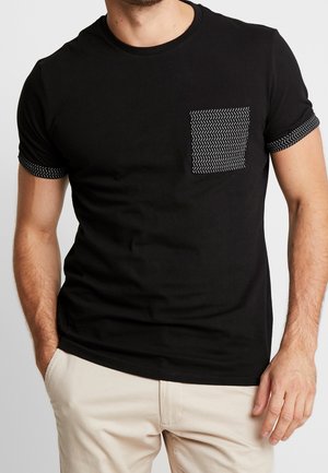 Mann trägt ein schwarzes kurzärmeliges T-Shirt mit gemusterten Bündchen und passender gemusterter Brusttasche, kombiniert mit hellbeigen Hosen.