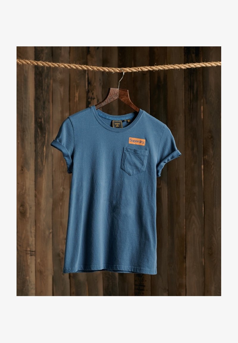 Modrá bavlněná tričko s krátkými rukávy, kulatým výstřihem, kapsou na levé straně a oranžovou nášivkou "Superdry" na hrudi.
