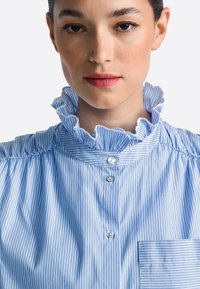 Blue and white striped shirt with a ruffled collar, four buttons, and a small pocket. Smooth fabric with a light texture.