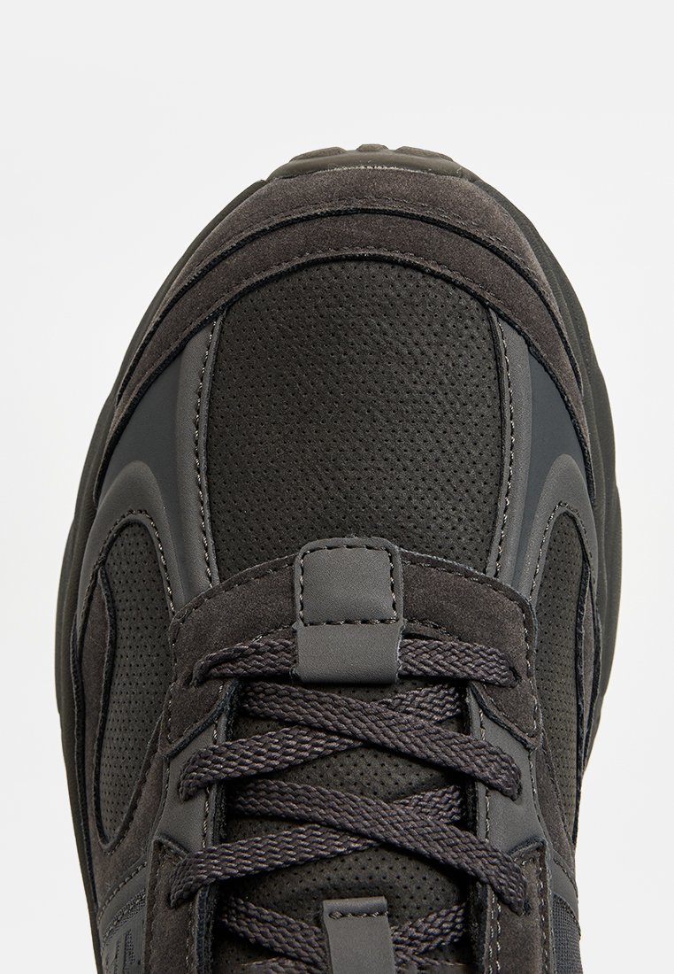 Zapatilla deportiva negra con parte superior de cuero texturizado y malla, con puntera reforzada, acentos grises y cordones. Diseño de suela plana.