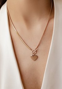 Collier en métal rose doré avec une chaîne torsadée, comportant un fermoir circulaire et un pendentif en forme de cœur comme détail clé.