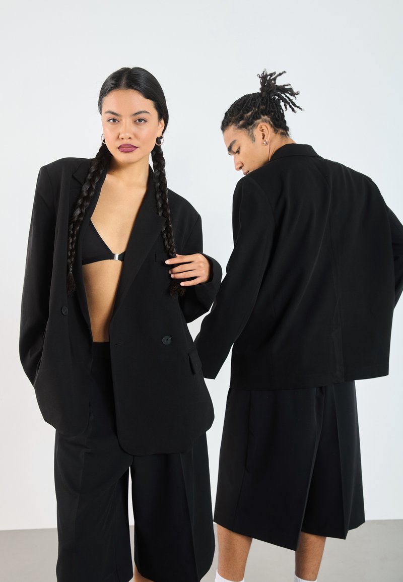 Black oversized blazers worn with matching shorts. The fabric is smooth, featuring a relaxed fit and double-button closure.