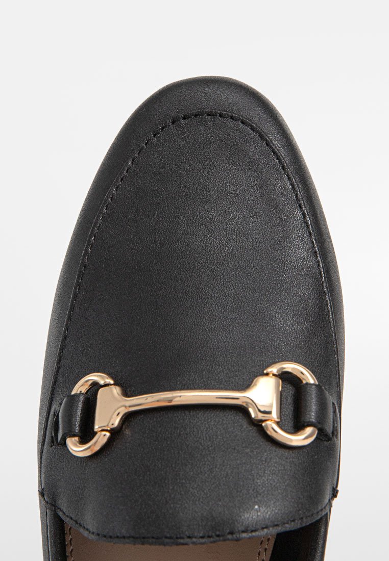 Mocasin de cuero negro con punta redondeada, que presenta un acento metálico dorado en la parte superior y una textura suave.