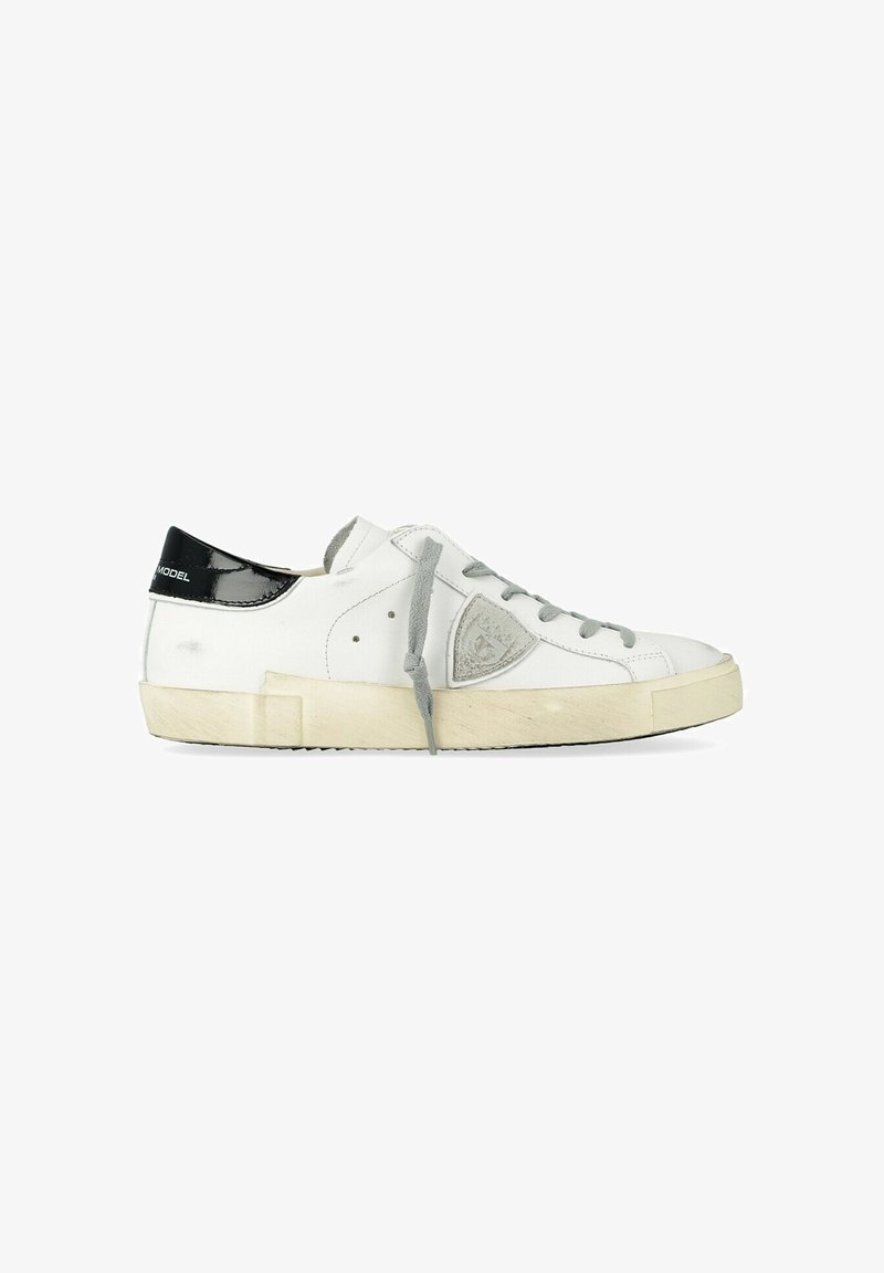 Baskets basses blanches avec des lacets gris, une semelle beige, une languette de talon noire marquée « MODEL » et un emblème gris sur le côté.