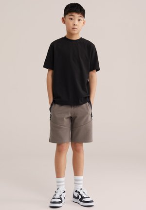 Fiú áll zsebre tett kézzel, fekete pólót, szürke rövidnadrágot, fehér zoknit és fekete-fehér tornacipőt visel, egyszínű háttér előtt.