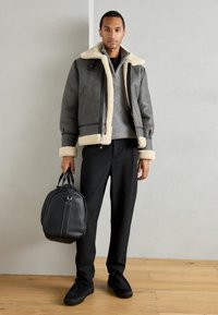Γκρι μπουφάν Shearling με κρέμα επένδυση, μαύρο πουλόβερ, μαύρο παντελόνι και μαύρη τσάντα σε έναν σύγχρονο εσωτερικό χώρο με ξύλινο δάπεδο.