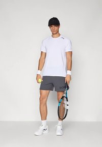 T-shirt branca de manga curta, calções cinzentos com estampa "BORD", sapatilhas brancas e boné preto. Segurando uma raquete e bola de ténis.