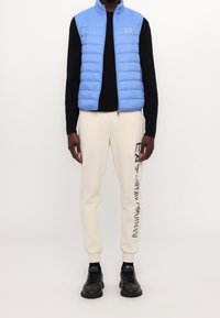 Světle modrá péřová vesta se zapínáním na zip a stojáčkem, spárovaná s krémovými tepláky s černým logem, a černými teniskami.