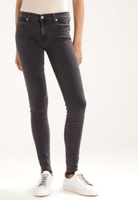 Mujer vestida con jeans negros ajustados y zapatillas blancas, de pie frente a un fondo claro, recortada a la altura de la cintura.
