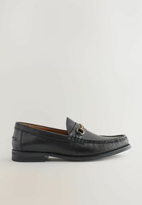 Mocasín de cuero negro con superficie texturizada, puntera redonda, acento metálico decorativo y una suela baja de goma. Diseñado para un fácil uso al deslizarse.