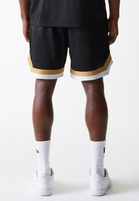 Svarte mesh basketballshorts med gull- og hvite detaljer, med elastisk midjebånd og en avslappet passform. Brukt sammen med hvite joggesko og sokker.