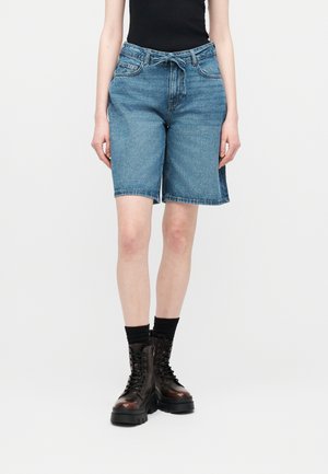 Person iført blå knælange denimshorts med et bundet bælte, sort ærmeløs top, sorte sokker og mørkebrune snørebåndsstøvler.