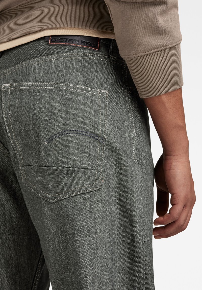 Achteraanzicht van een persoon die grijze denim jeans draagt met gestikte achterzakken en een beige langemouwen top met geribbelde manchetten.