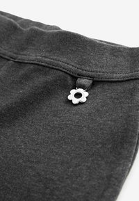 Graue Jogginghosen aus einem glatten Material, mit einem floralen silbernen Metallakzent am Bund. Sichtbare Nähte entlang der Kanten.