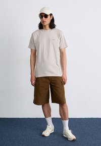 En ung mann står mot en ensfarget vegg, iført en lys beige t-skjorte, brune cargo-shorts, en hvit caps og solbriller, med hvite sokker og sneakers.
