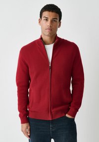 Man wearing a red zip-up sweater over a white shirt and dark jeans, standing against a plain light background with one hand in pocket.