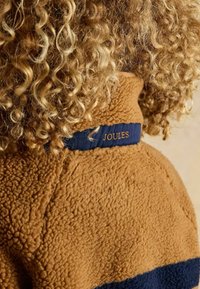 Izmēra tonētu fleecēta jaka ar teksturētu virsmu, kurai ir tumši zilas krāsas josla ap apkakli, uz kuras ir izšauts "JOULES" zelta krāsā.