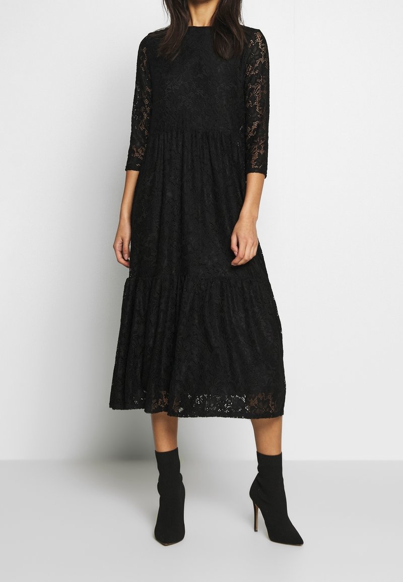 Femme portant une robe midi en dentelle noire à manches trois-quarts et des bottines noires à talons hauts, debout devant un fond blanc.