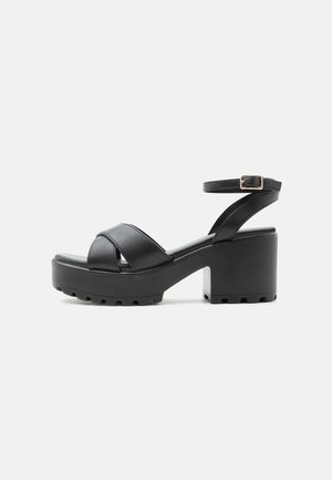 Sort sandal med chunky hæl, krydsede stropper og justerbar ankelrem med spænde på en profileret platformsål.